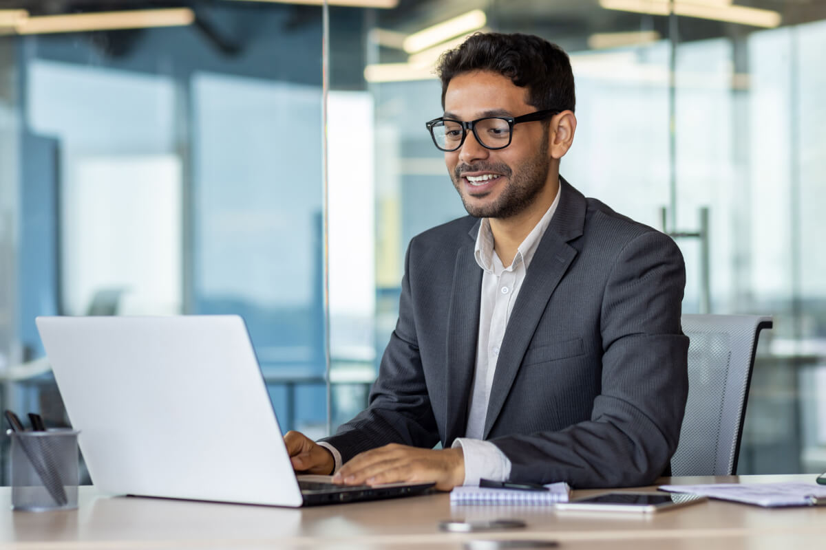 Male in a suit and black glasses sits in a corporate office conference room while working on a laptop to decide on the right merchant services provider for his company